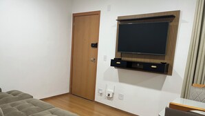 A modern living room with a flat-screen TV mounted on a wooden panel, a grey sofa, and a wooden door. - Apartment 15 minutes from Confins International Airport (Lagoa Santa)