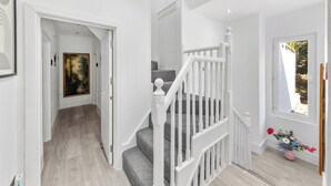 Eine Treppe mit weißen Geländern führt zu einer oberen Etage, eine Tür mit grünem Ausgangsschild und ein Fenster mit Blick auf Grünflächen.