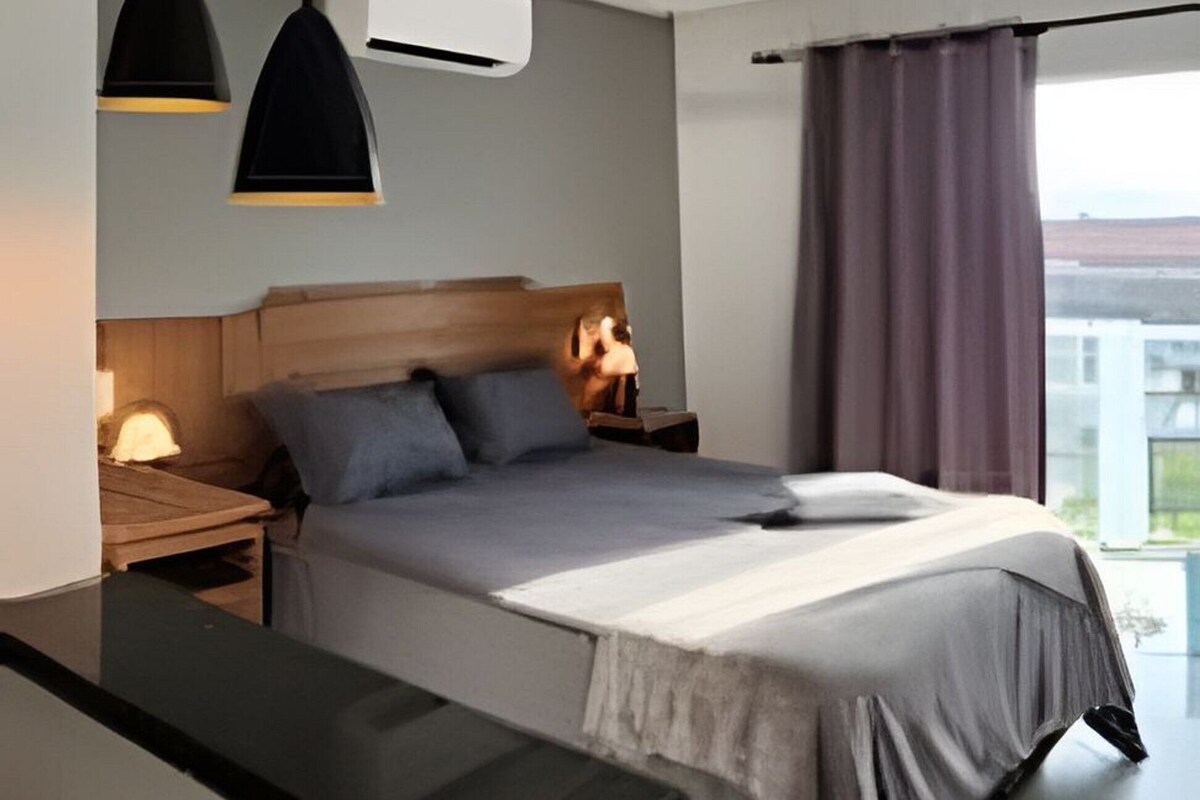 Un dormitorio moderno con cama, mesitas de noche, un escritorio, lámparas colgantes y vistas a través de una ventana.