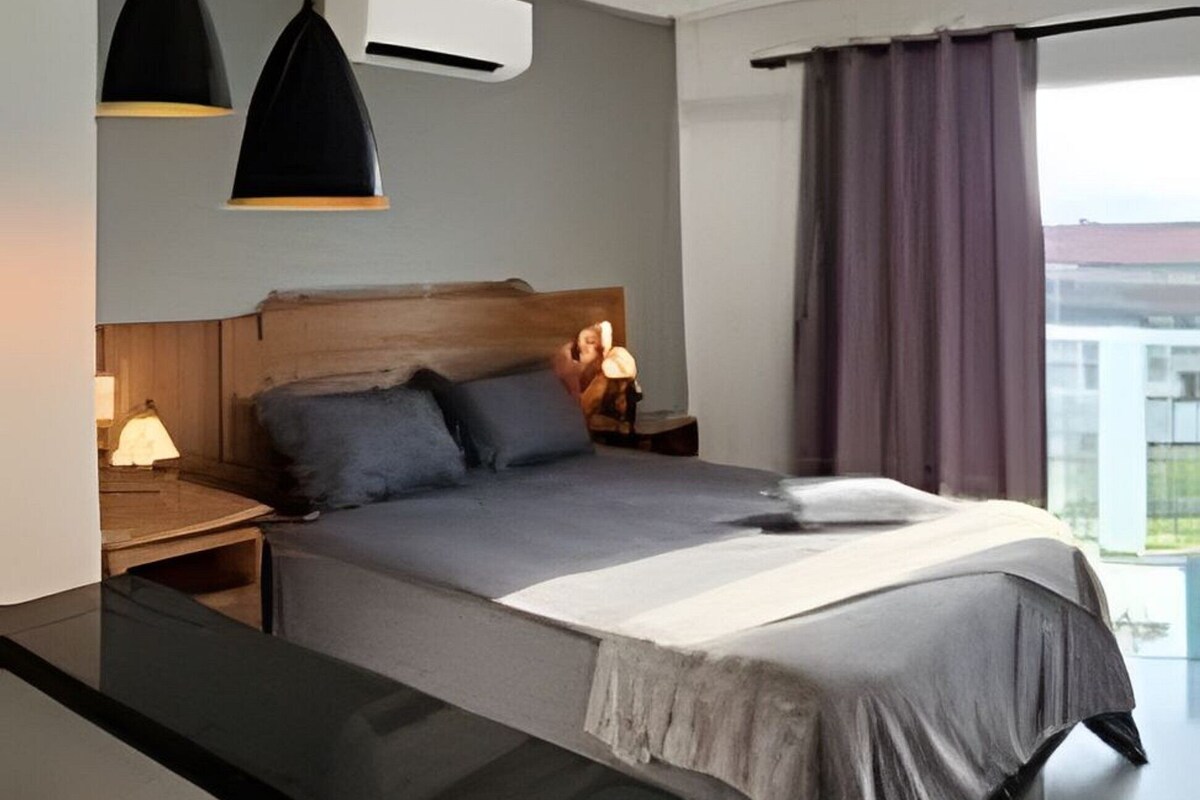 Un dormitorio moderno con cama, mesitas de noche, un escritorio, lámparas colgantes y una ventana con cortinas.