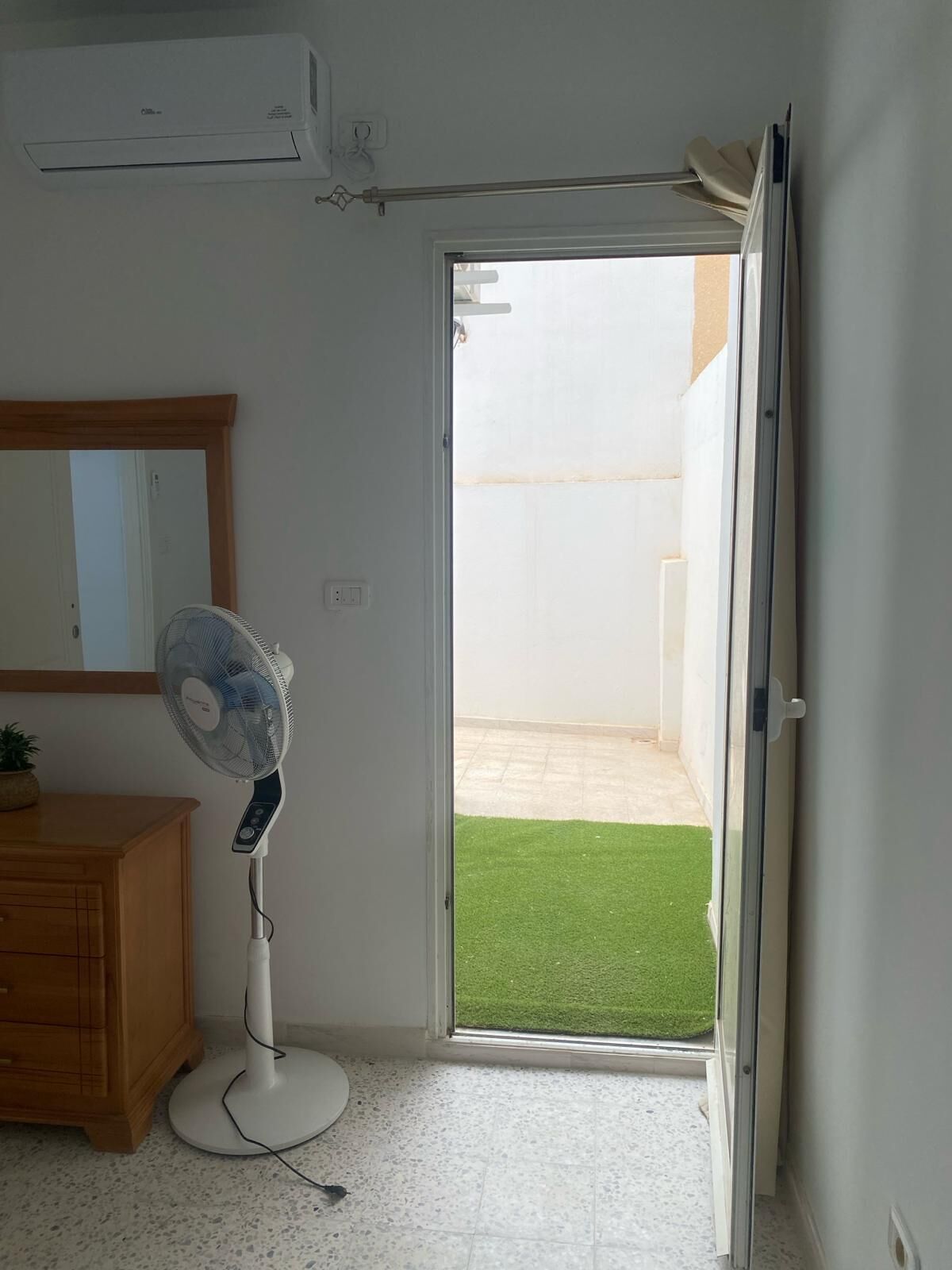 Habitació amb porta de vidre corredissa que dona a un balcó amb gespa artificial, un ventilador de peu i un armari de fusta.