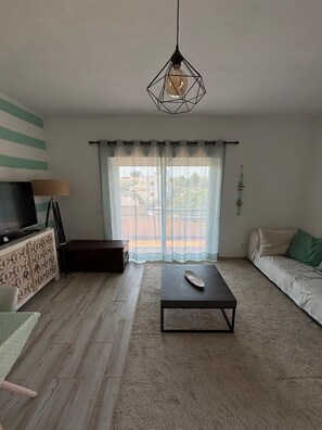 A living room with a sofa, a coffee table, a TV on a stand, a patterned cabinet, and a view through a window with curtains. - The Riscas House—Apartment in Alvor Brisamar (portimao)