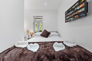 A modern hotel room with a large bed, a wall-mounted TV displaying movie options, a window with blinds, and a bedside table with a lamp. - Oak Lane Studio (London)