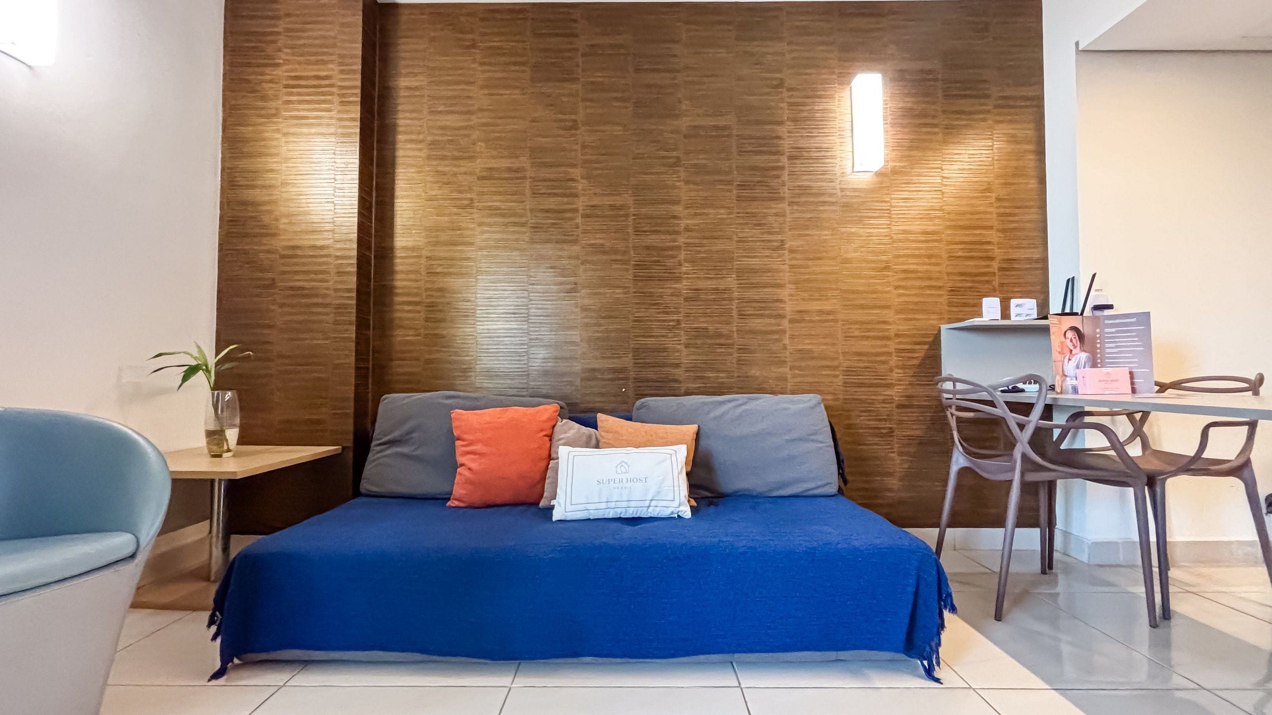 A modern hotel room with a blue bed, a small table with a plant, and a desk with a chair.