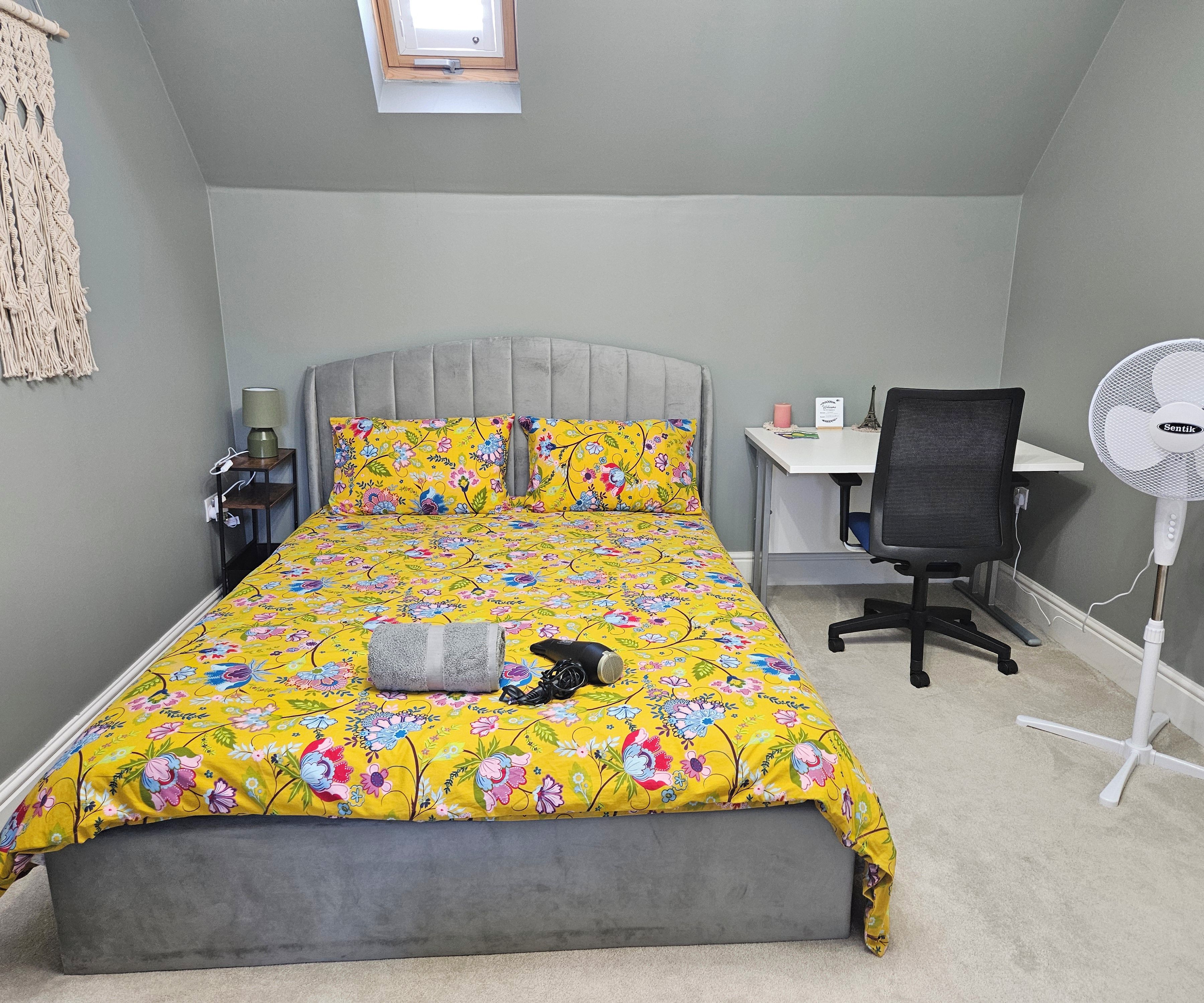 A bedroom with a bed, a desk, a chair, a standing fan, and a small window.
