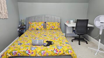 A bedroom with a bed, a desk, a chair, a standing fan, and a small window.