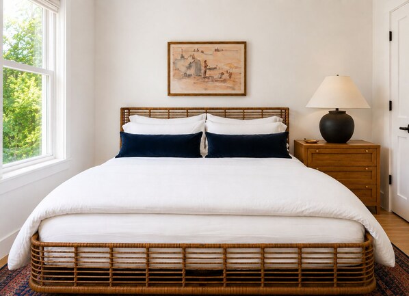 Uma cama com roupa de cama branca e azul-marinho, cabeceira de madeira, mesa de cabeceira com abajur e um quadro na parede.