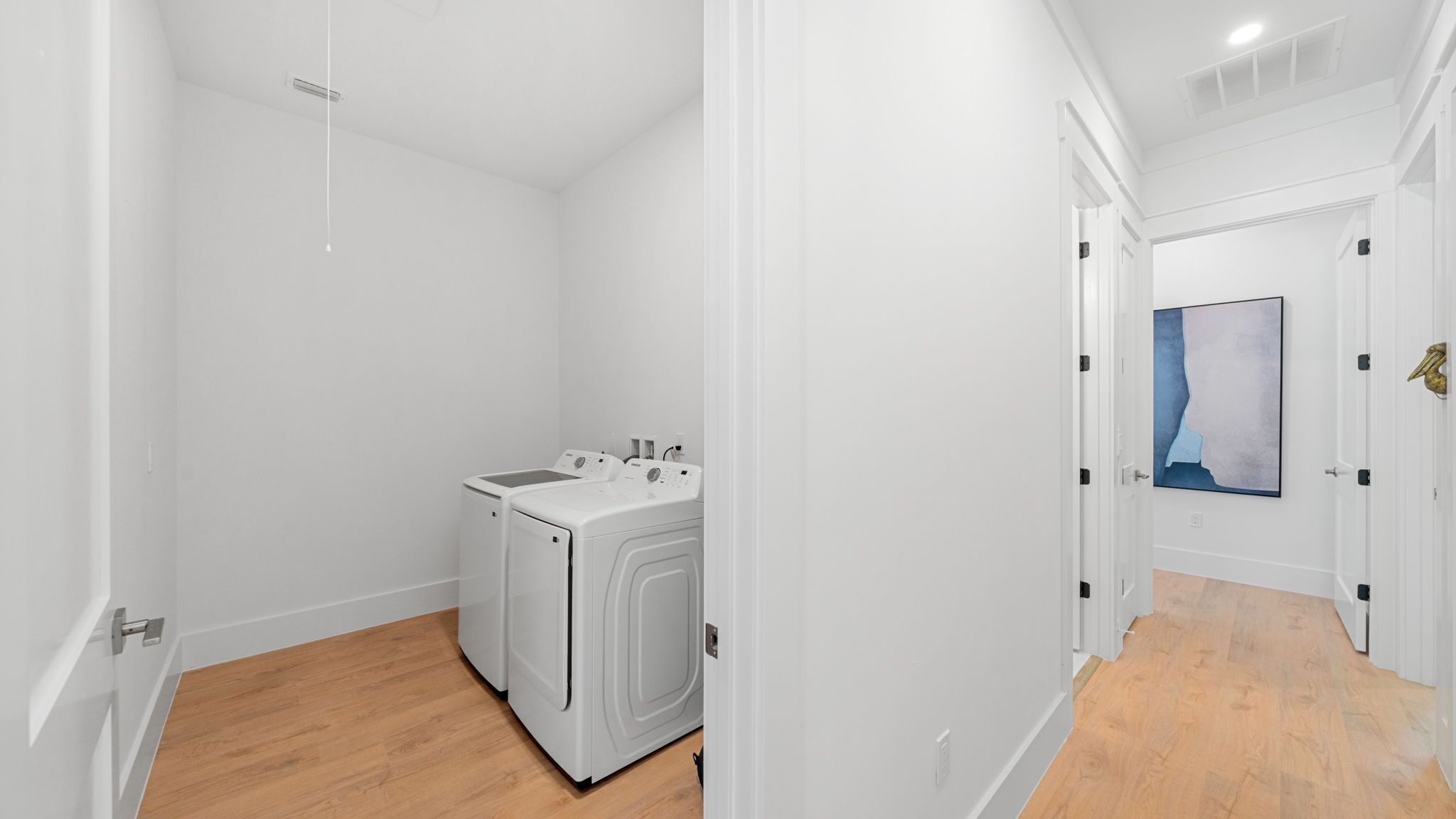 Un cuarto de lavandería con lavadora y secadora, un pasillo con piso de madera y una puerta con un cuadro.