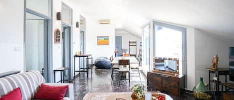 A spacious living area with a sofa, coffee table, and a dining table with chairs. There is a large mirror above the dining table and a window providing natural light.