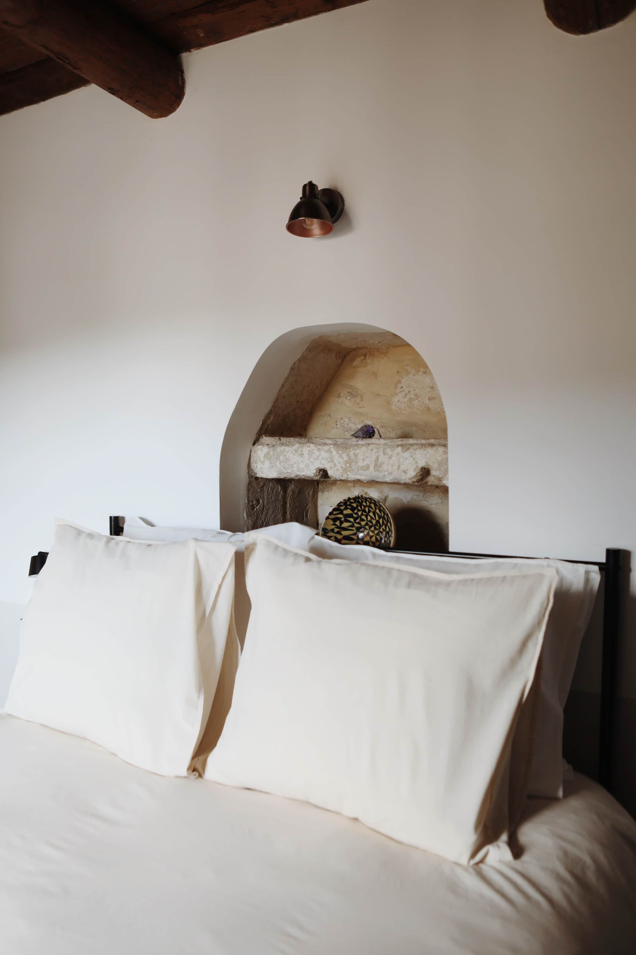 A bed with white linens, a stone archway, and a small shelf with decorative items.