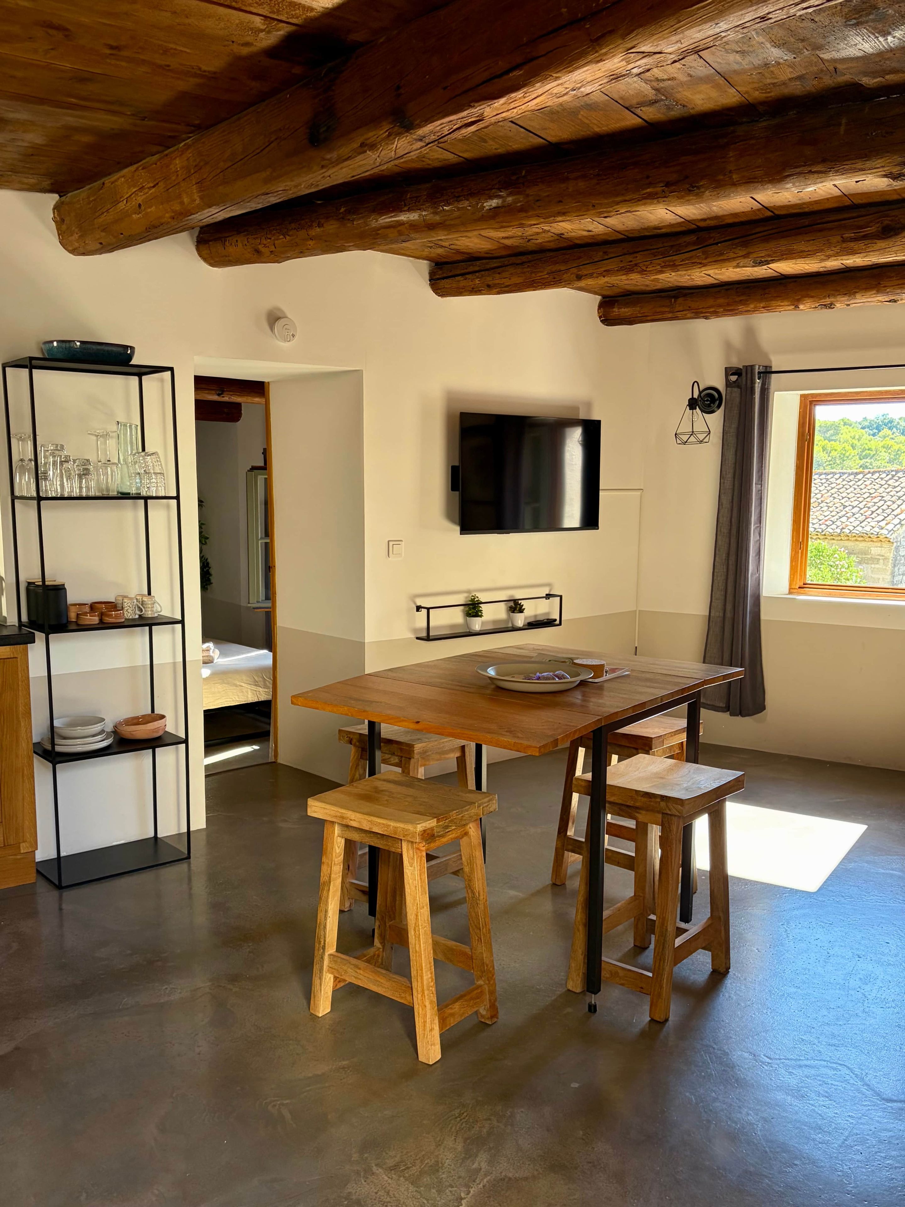 Une pièce avec une table en bois, deux tabourets, un téléviseur fixé au mur et une fenêtre avec des rideaux.