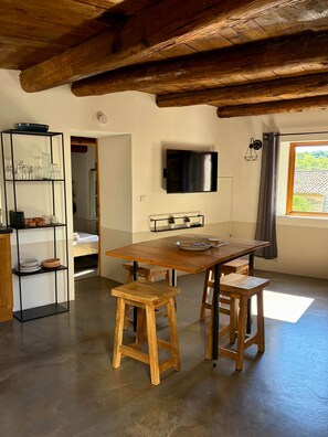 A room with a wooden table, two stools, a TV mounted on the wall, and a window with curtains. - La Vallete Boutique - La Petite Valette (Sanilhac-Sagriès)