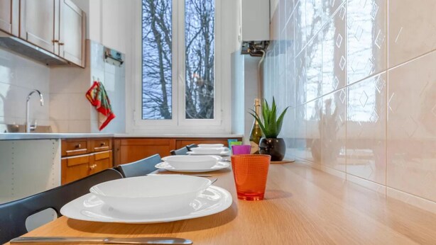 Ett modernt kök med ett matbord för fyra personer, ett fönster med utsikt över träd och en krukväxt på köksbänken.