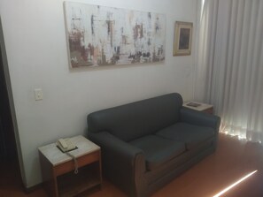 A living room with a dark-colored sofa, a small wooden side table with a telephone, a wall-mounted abstract painting, and a framed picture. - LIBERDADE SQUARE SAVASSI/LOURDES (Belo Horizonte)