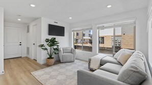 Ein modernes Wohnzimmer mit einem grauen Sofa, einem Topf mit einer Pflanze und einem großen Fenster mit Blick auf eine Baustelle.