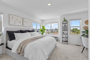 A bedroom with a large bed, a window with a view, a plant, and a shelf with decorative items. - Brand-New 3-Bedroom Townhome Near Golden, Boulder & Ralston Creek Trail. (Arvada)