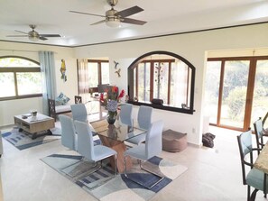 A modern dining area with a glass table, light blue chairs, and a ceiling fan. - Flor de Palma Villa (Tronadora)