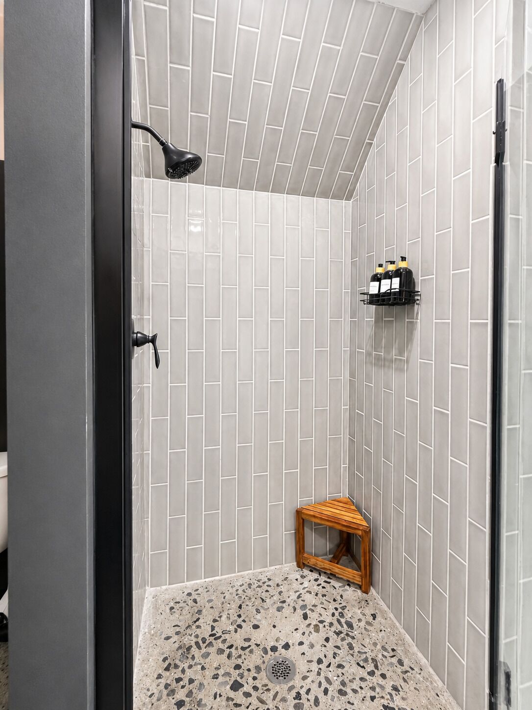 A shower with a tiled wall, a wooden stool, and a shelf with bottles.