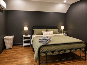 A bedroom with a metal bed frame, a green bedspread, two bedside tables with lamps, a small plant, and a laundry basket.