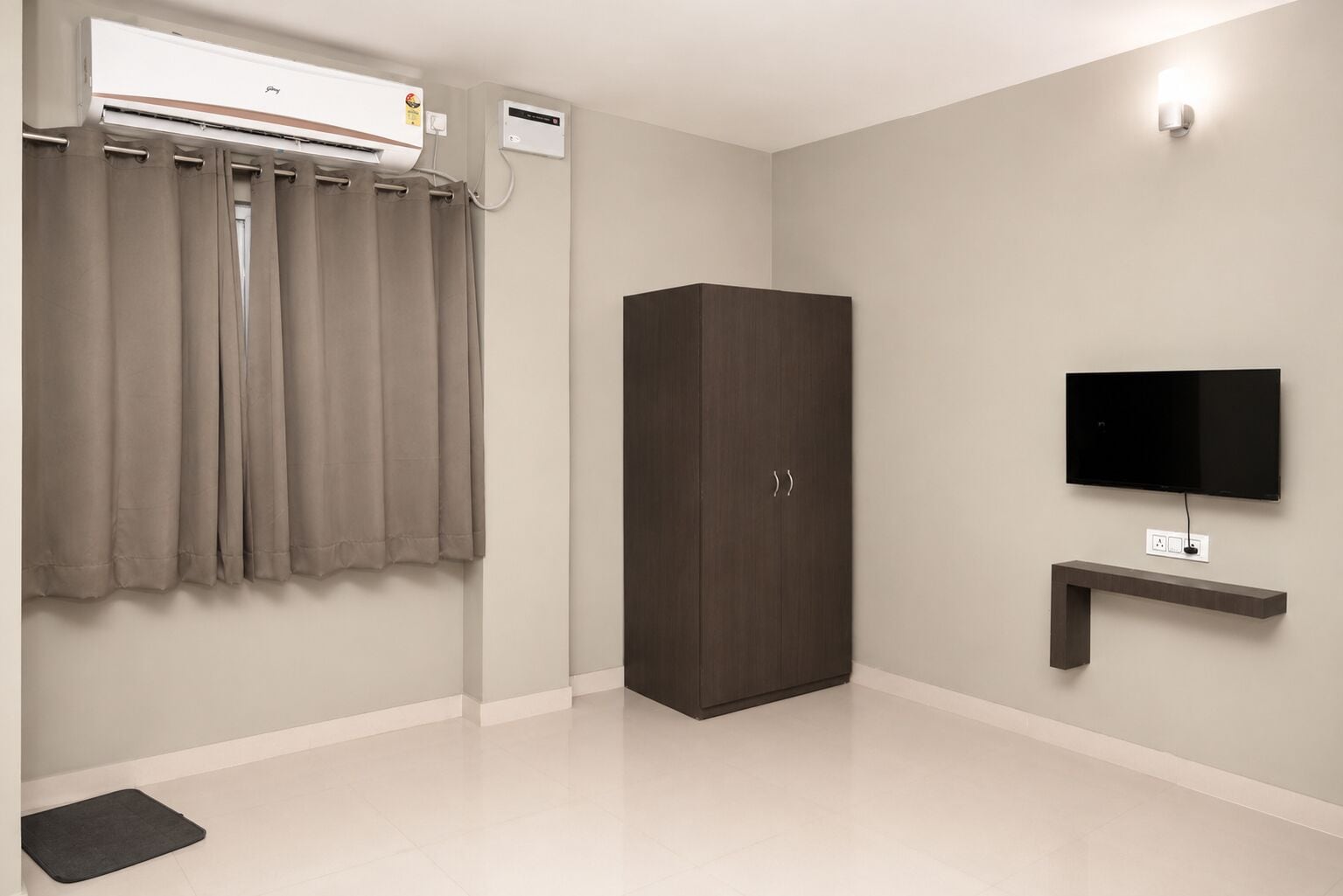 A room with a wardrobe, a flat-screen TV mounted on the wall, a ceiling-mounted air conditioner, and a small shelf below the TV.