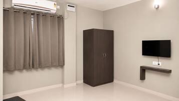 A room with a wardrobe, a flat-screen TV mounted on the wall, a ceiling-mounted air conditioner, and a small shelf below the TV.