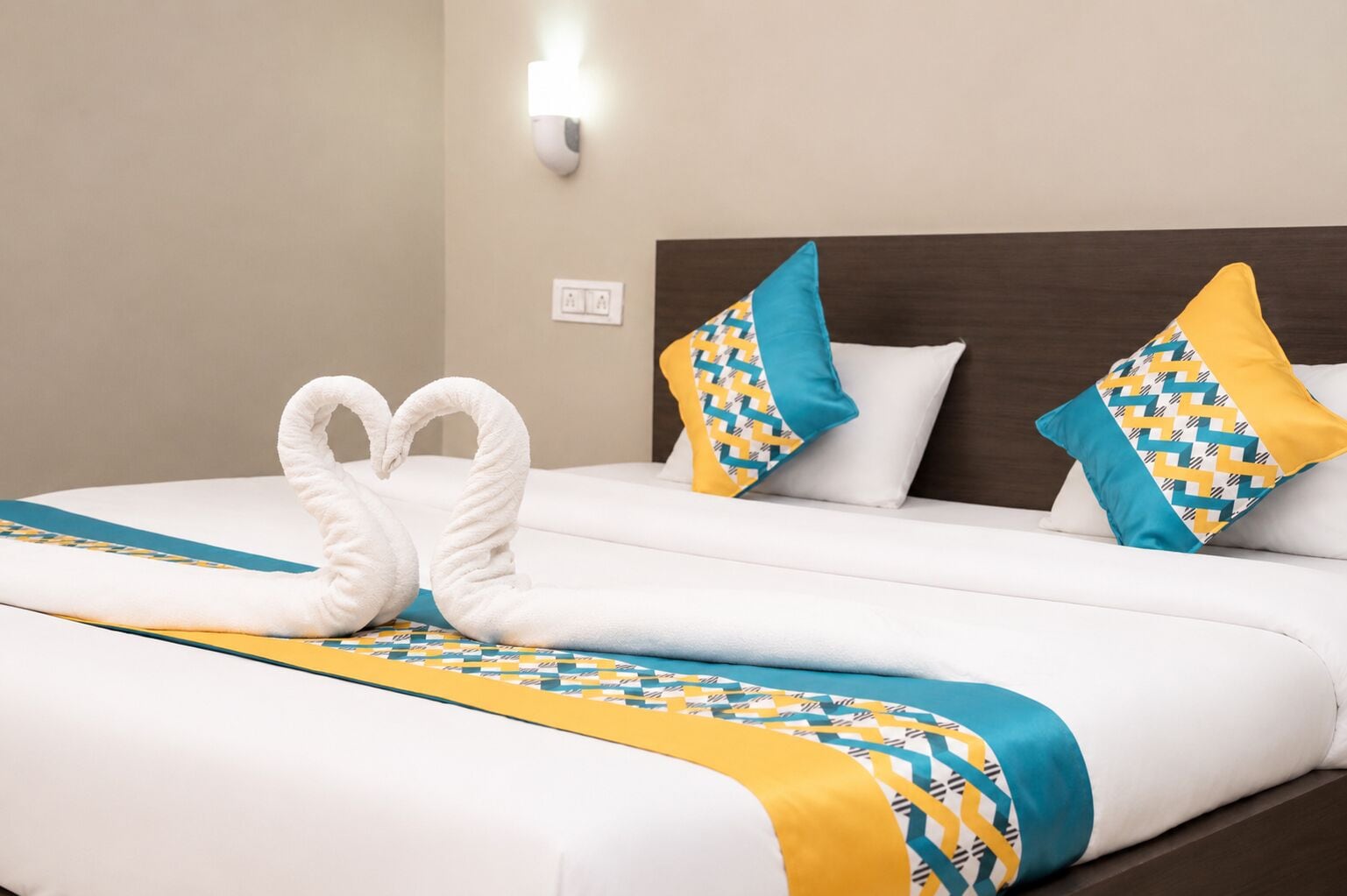 A neatly made bed with a white and yellow patterned bedspread, two pillows with a blue and yellow geometric design, and two white towels arranged in a heart shape.