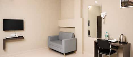 Habitación de hotel con televisor de pantalla plana, un sofá gris, un escritorio con silla, un espejo y un cuadro en la pared.