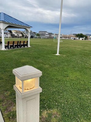 Un champ verdoyant avec un kiosque, des bancs et un grand poteau blanc.