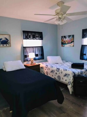 A room with two beds, a ceiling fan, a nightstand with a lamp, and a painting on the wall.
