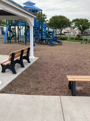A playground with a slide and climbing structures, surrounded by mulch, benches, and trees.