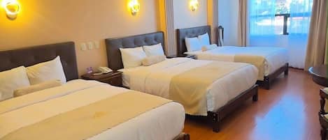 A hotel room with three beds, wooden flooring, and wall-mounted lighting.