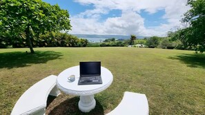 Ein Laptop auf einem weißen Tisch, daneben eine Tasse, platziert auf einer Rasenfläche mit Blick auf eine Landschaft.