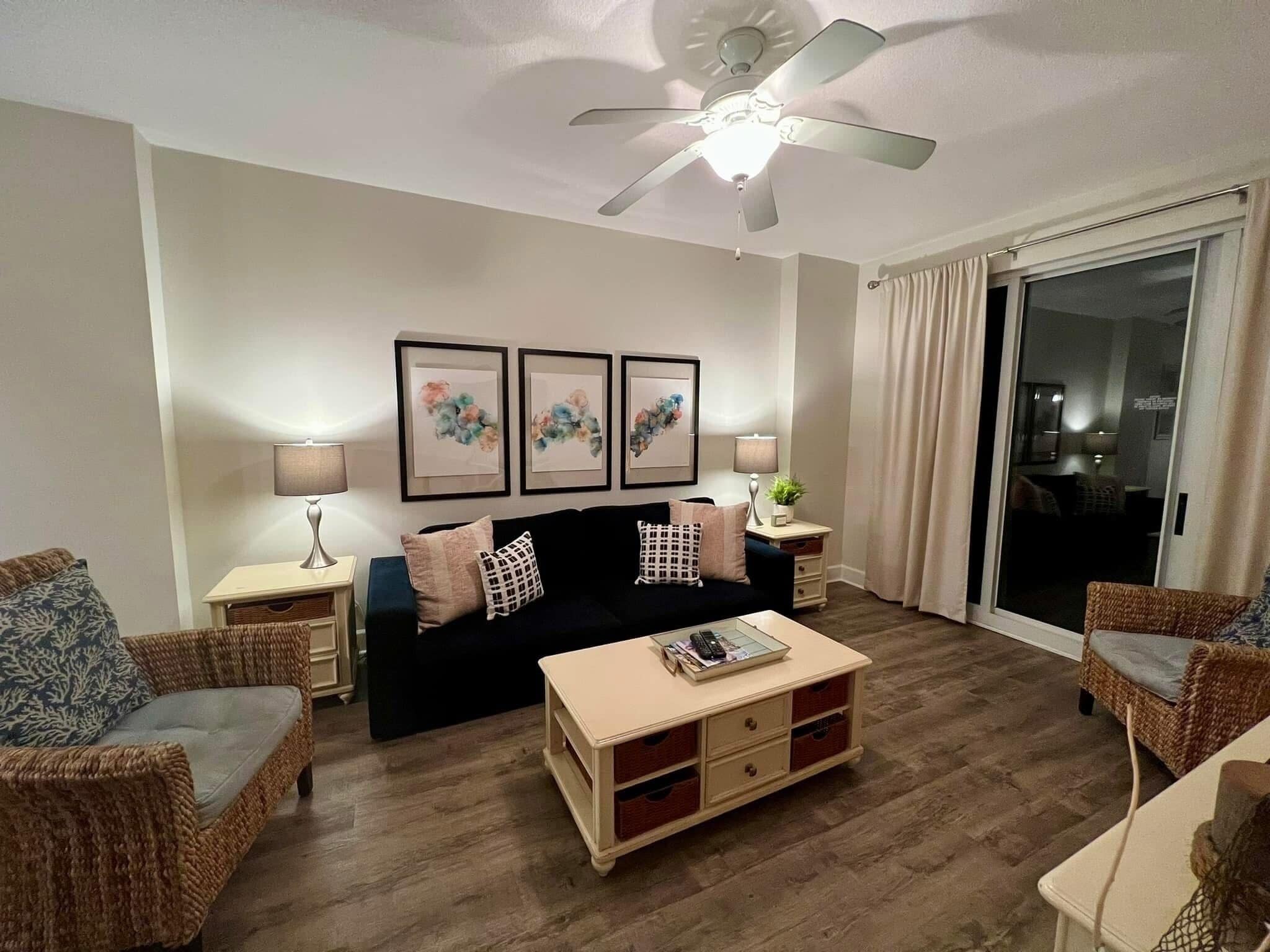 A living room with a black sofa, two side tables with lamps, a ceiling fan, and a sliding glass door.