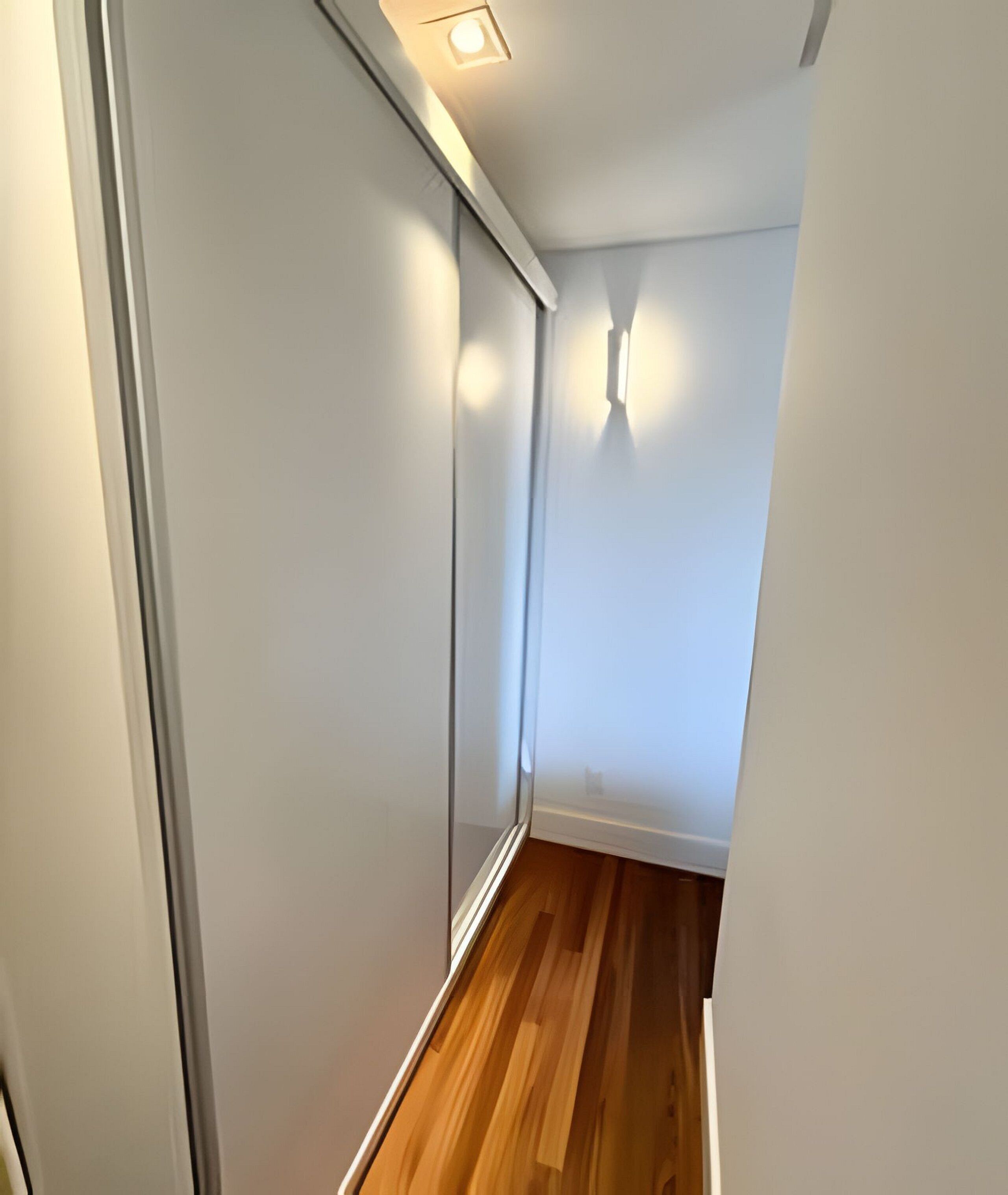 A narrow hallway with a wooden floor, white walls, and a ceiling light fixture.