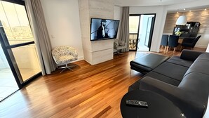 Ett modernt vardagsrum med en stor tv-skärm, en svart lädersoffa, en mönstrad fåtölj och ett trägolv.