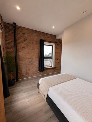 A bedroom with a brick wall, a window with curtains, a bed with a white bedspread, and a potted plant. - Boutique Suite in Historic Downtown Renfrew (Renfrew)