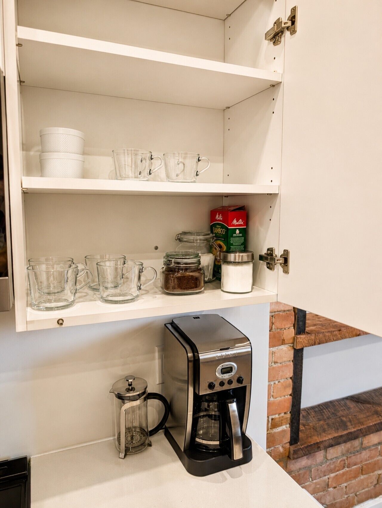 A kitchen with a coffee maker, glass mugs, and a box of instant coffee.