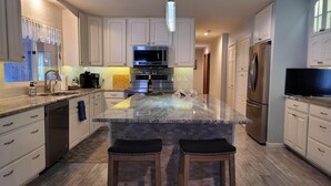 A modern kitchen with white cabinetry, a central island, stainless steel appliances, and a flat-screen TV. - Luxury Home with Indoor Pool, 3-bedroom house in delightful Milford MI (Milford)
