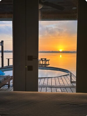 Vue depuis une pièce donnant sur une terrasse en bois, qui surplombe une étendue d’eau au coucher du soleil.