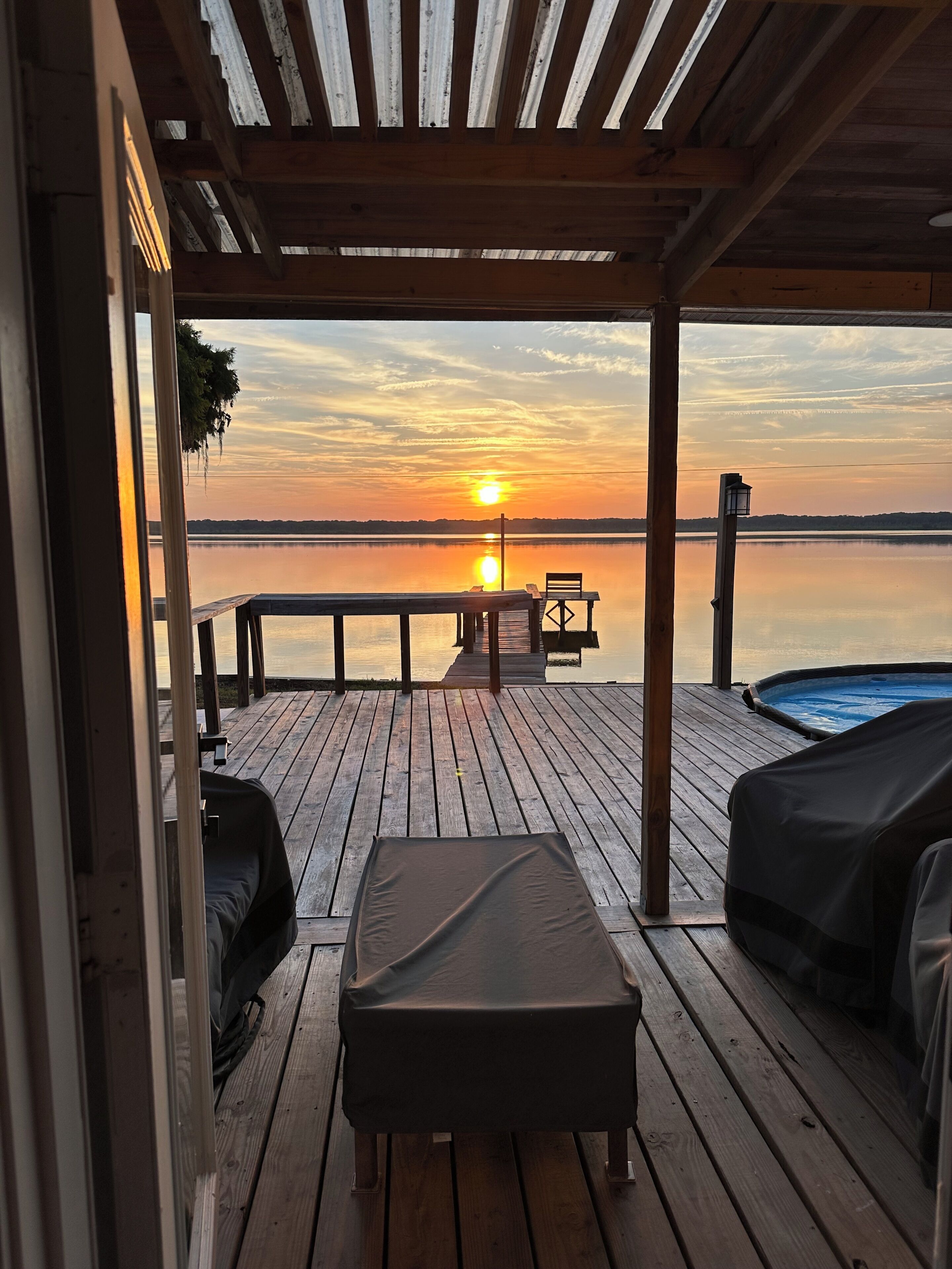 A wooden deck with a bench and a hot tub, overlooking a calm body of water during sunset.