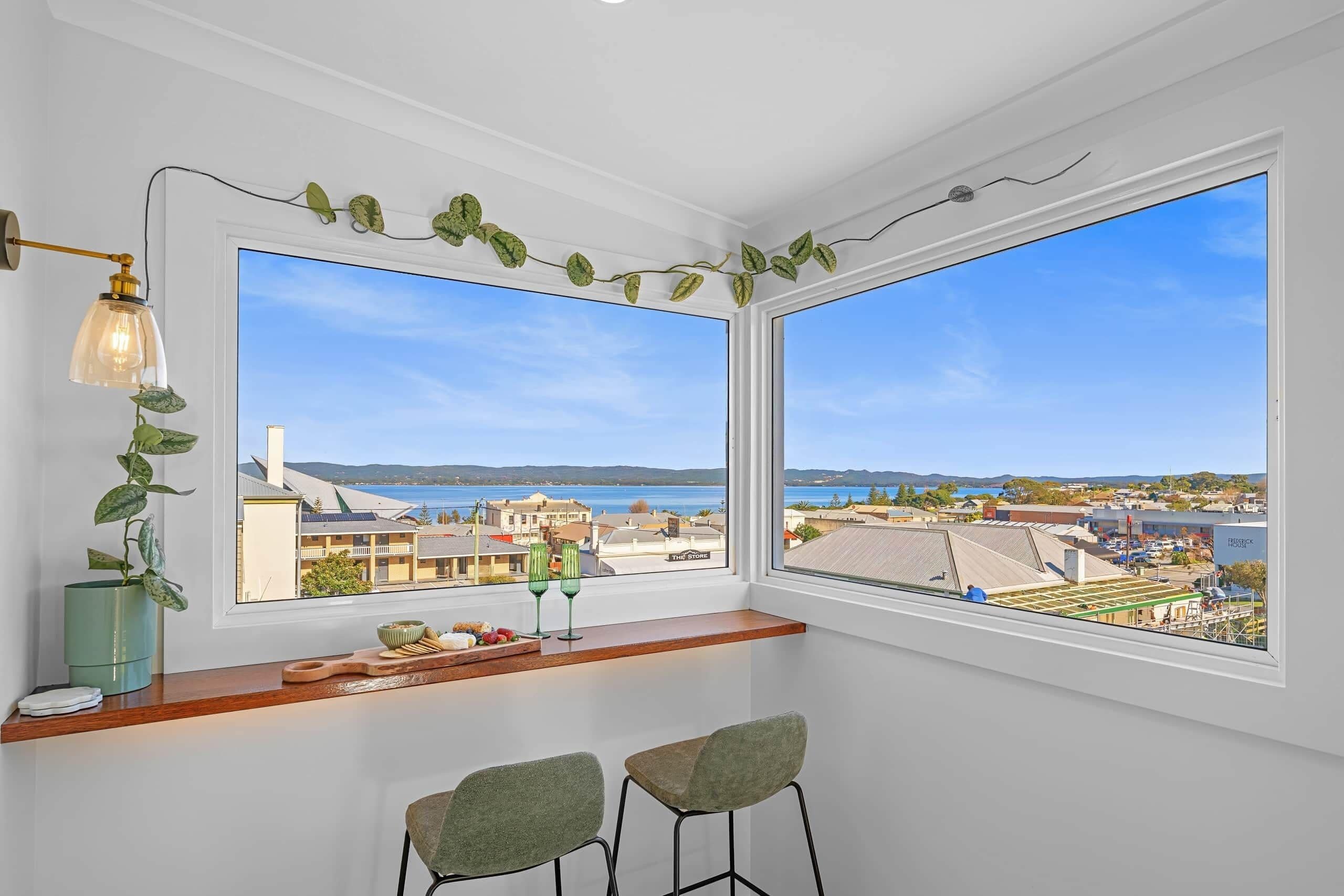 A room with two large windows overlooking a coastal town, a small bar area with stools, and a decorative plant garland.
