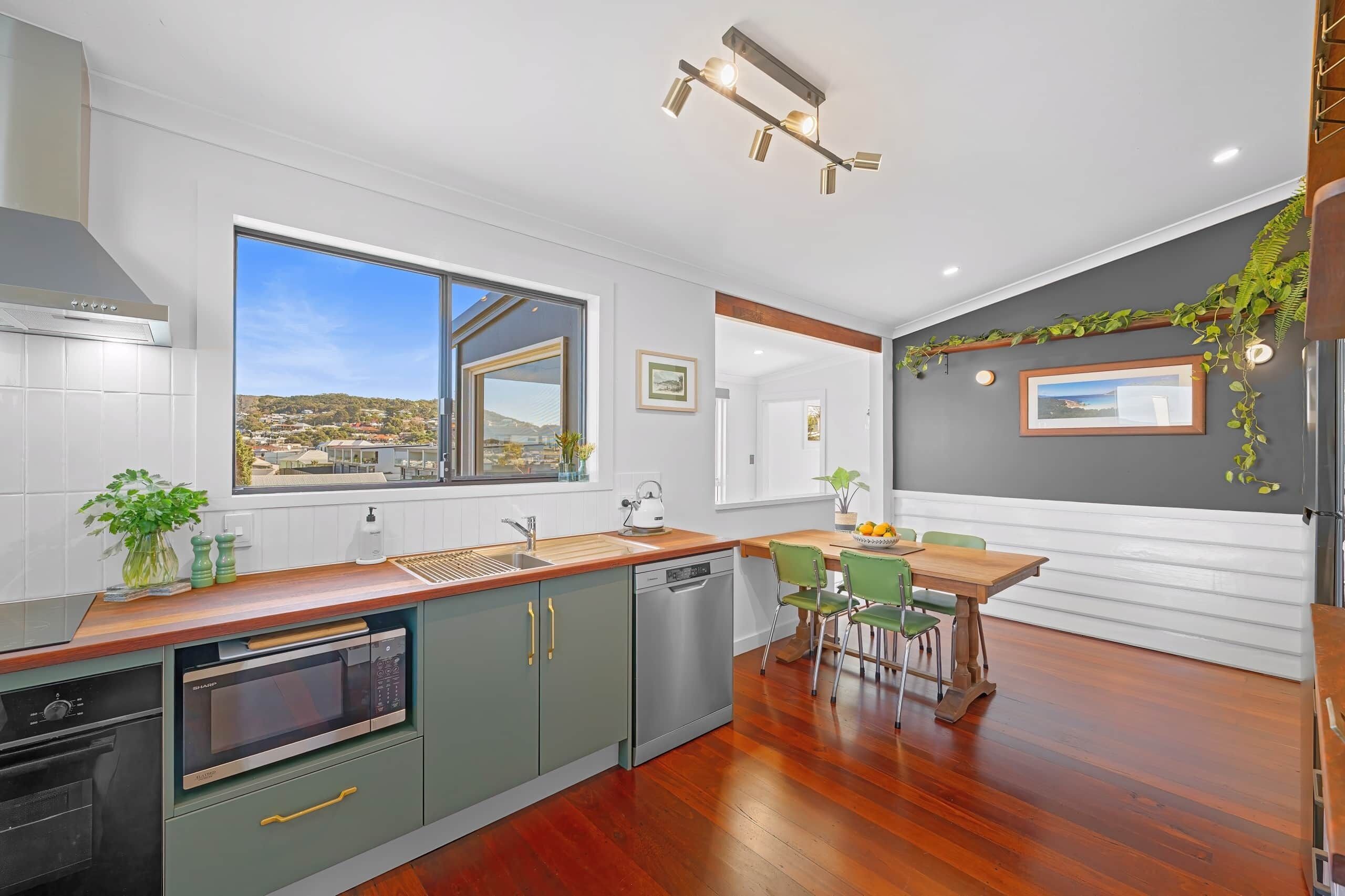 A modern kitchen with a large window, a dining area with a wooden table and chairs, and a view of a cityscape.