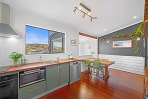 A modern kitchen with a large window, a dining area with a wooden table and chairs, and a view of a cityscape. - The Yellow Door on Spencer | Harbour Views (Albany)