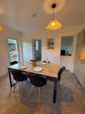 A dining area with a wooden table, four chairs, a clear plastic table cover, a bottle, and a framed picture on the wall. - Cozy House (Saumur)
