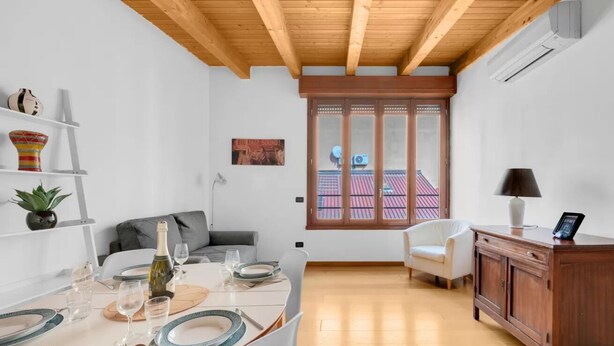 Zona pranzo con tavolo apparecchiato per quattro persone, divano, credenza e vista su un edificio con tetti rossi attraverso una porta vetrata.