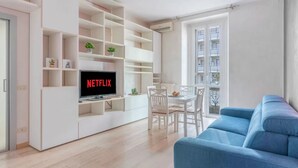 Uma sala de estar moderna com televisão de tela plana, estantes brancas, um sofá azul e um espaço de jantar com cadeiras brancas.