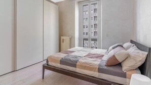 Quarto com uma cama, radiador, janela com vista para edifícios e guarda-roupa.