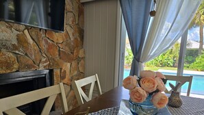 A stone wall, a fireplace, a dining table with chairs, a vase with flowers, and a view of a swimming pool. - Stylish Pool Retreat in the Heart of Vero Beach. (Vero Beach)