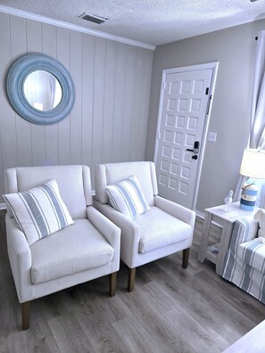 Ein Zimmer mit zwei weißen Sesseln, einem runden Spiegel, einer Tür und einem kleinen Tisch mit einer Lampe.