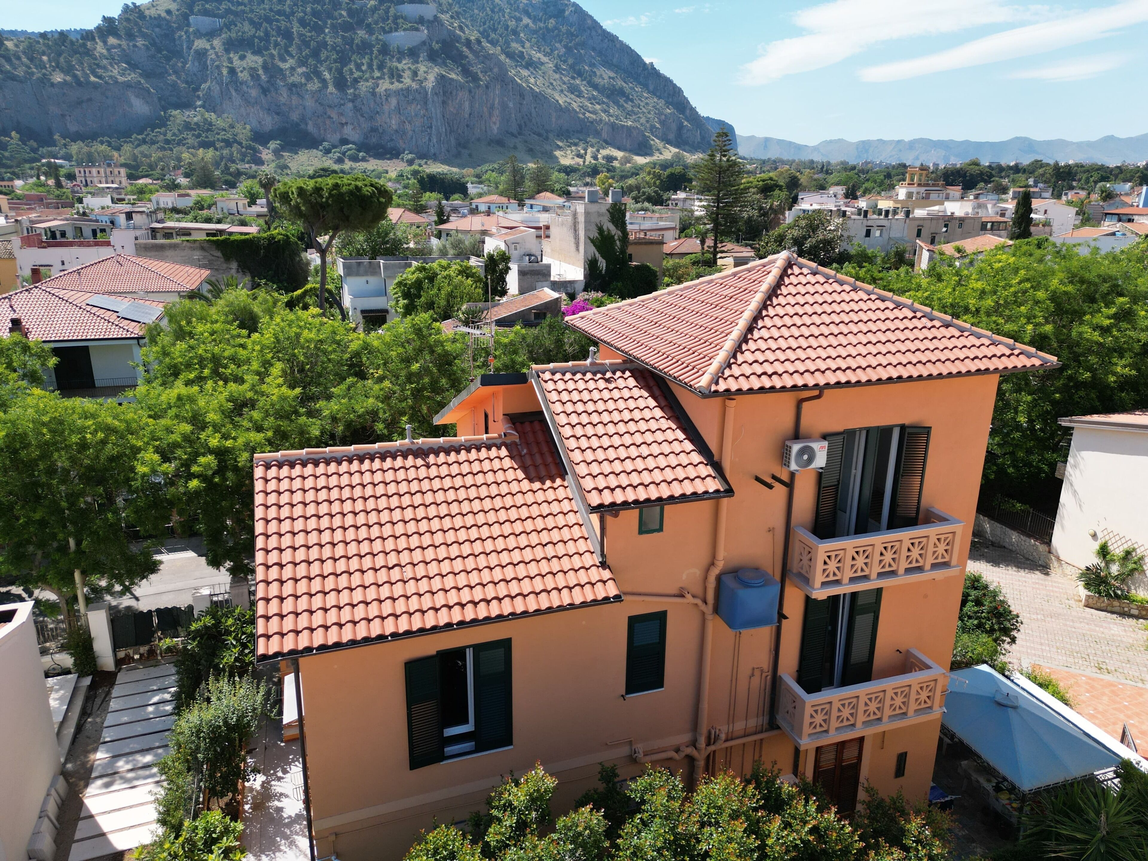 A multi-story building with a red-tiled roof, a balcony with a geometric design, and a view of a mountainous landscape in the background.